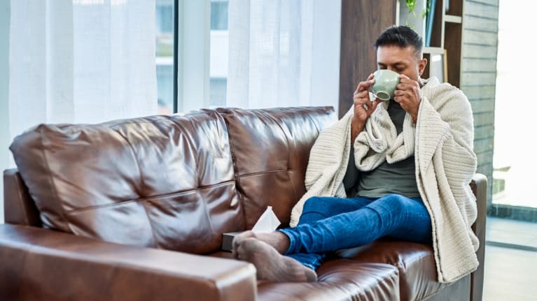 man having a warm drink while recovering from an illness on the sofa at home